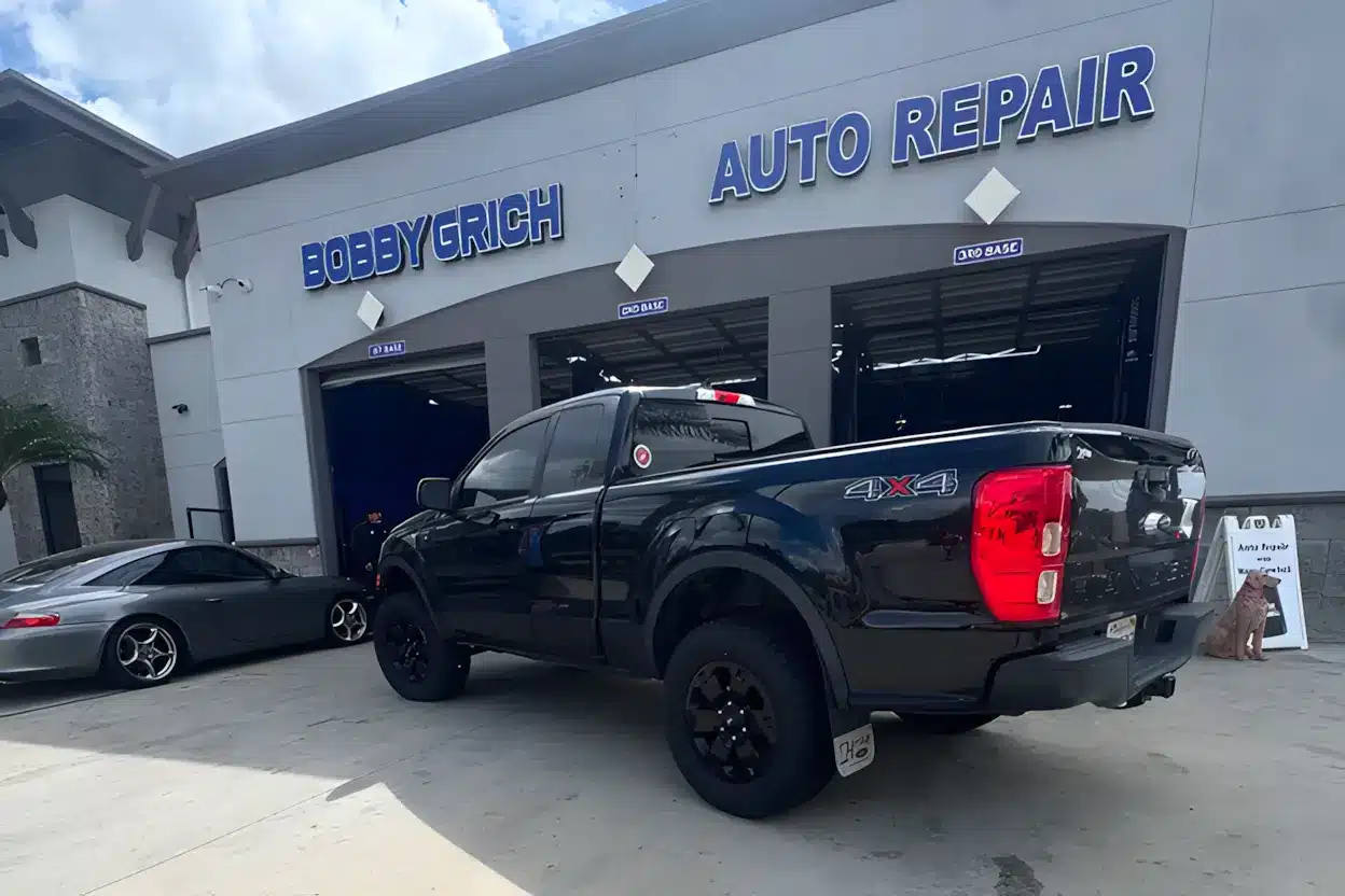 Black Ford Ranger 4x4 truck parked outside Bobby Grich Auto Repair, with service bays open and a gray Porsche in the background under a partly cloudy sky.