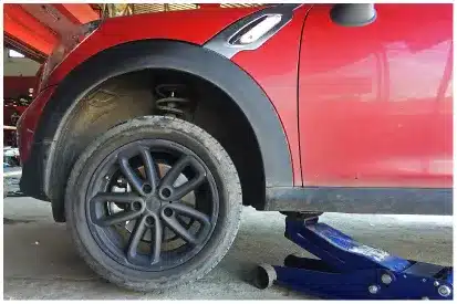 Tire Services In Rancho Santa Margarita, CA At Bobby Grich Auto Repair. A red car is lifted with a blue hydraulic floor jack under the rear side. The rear wheel, tire, and suspension are visible, and the car appears to be undergoing maintenance or repair.