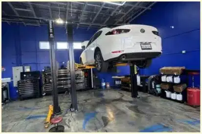 Vehicle Maintenance In Rancho Santa Margarita, CA At Bobby Grich Auto Repair. A white car is elevated on a hydraulic lift inside a clean, modern auto repair shop with blue walls, tool cabinets, and various supplies organized along the side.