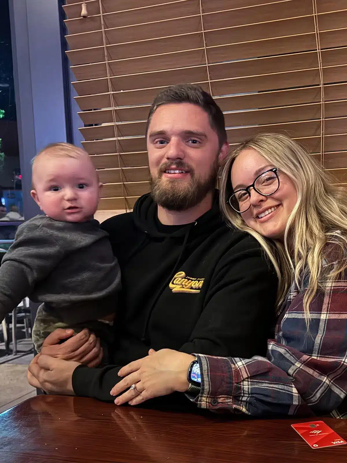 Josh Brady, Auto Shop Owner in In Rancho Santa Margarita, CA At Bobby Grich Auto Repair A man and woman sit closely together at a table, smiling at the camera. The man holds a baby on his lap. Behind them are wooden blinds, and they all appear happy and relaxed indoors.