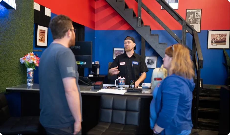 About Us, Auto Repair In Rancho Santa Margarita, CA At Bobby Grich Auto Repair. Two customers stand at a counter and talk with an employee in a black uniform inside a modern shop with blue and red walls, stairs, framed pictures, and a vase of flowers.