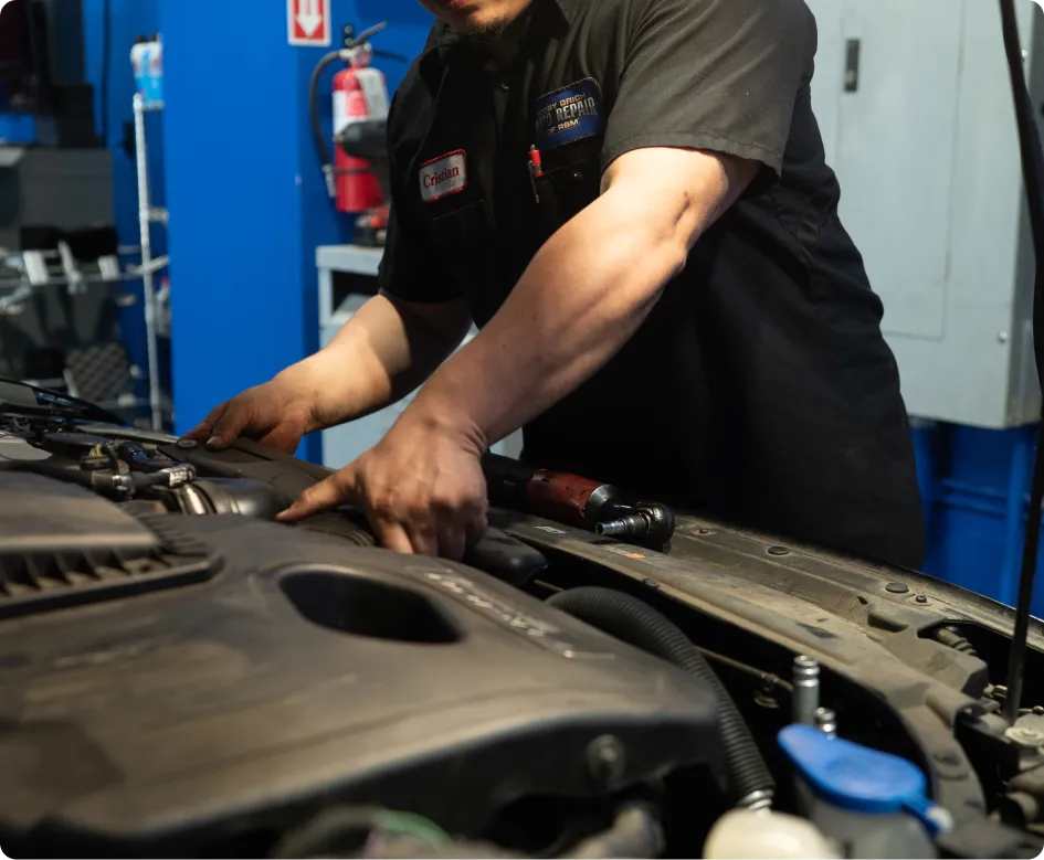 Audi Repair In Rancho Santa Margarita, CA At Bobby Grich Auto Repair. A mechanic wearing a black uniform works under the hood of a car in an auto repair shop. Tools and equipment are visible in the background.