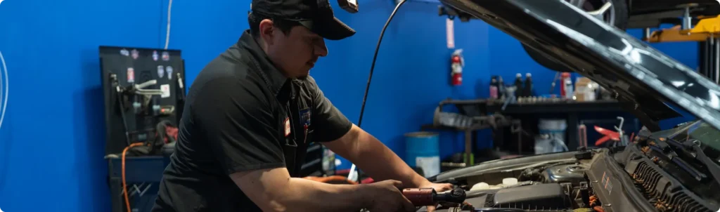 European Auto Repair In Rancho Santa Margarita, CA At Bobby Grich Auto Repair. A mechanic wearing a black uniform and cap uses a tool to work under the hood of a car in a garage with blue walls and various equipment in the background.
