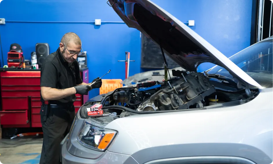 Fleet & Diesel Services In Rancho Santa Margarita, CA At Bobby Grich Auto Repair. A mechanic wearing gloves and safety glasses works on the engine of a silver SUV with the hood open in an auto repair shop with blue walls and red tool cabinets in the background.