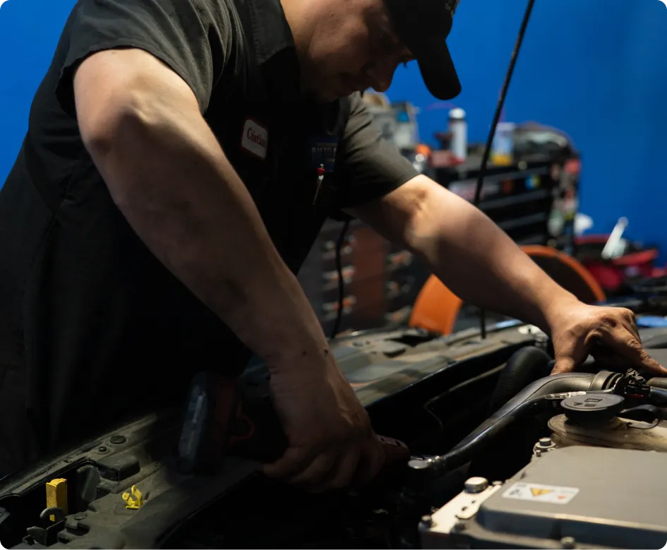 Land Rover Repair In Rancho Santa Margarita, CA At Bobby Grich Auto Repair. A mechanic wearing a black uniform works under the hood of a car, using a power tool. His arms are dirty, and automotive tools and equipment are visible in the background.