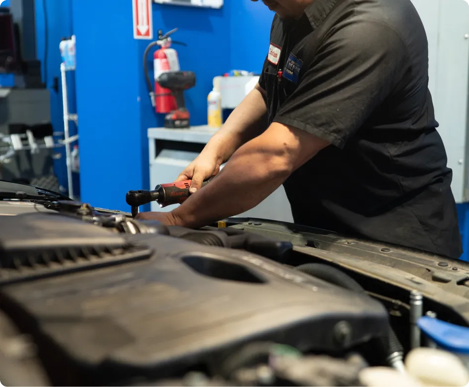Mercedes Repair In Rancho Santa Margarita, CA At Bobby Grich Auto Repair. A mechanic wearing a black uniform uses a power tool to work on a car engine in an auto repair shop, with various tools and a fire extinguisher visible in the background.