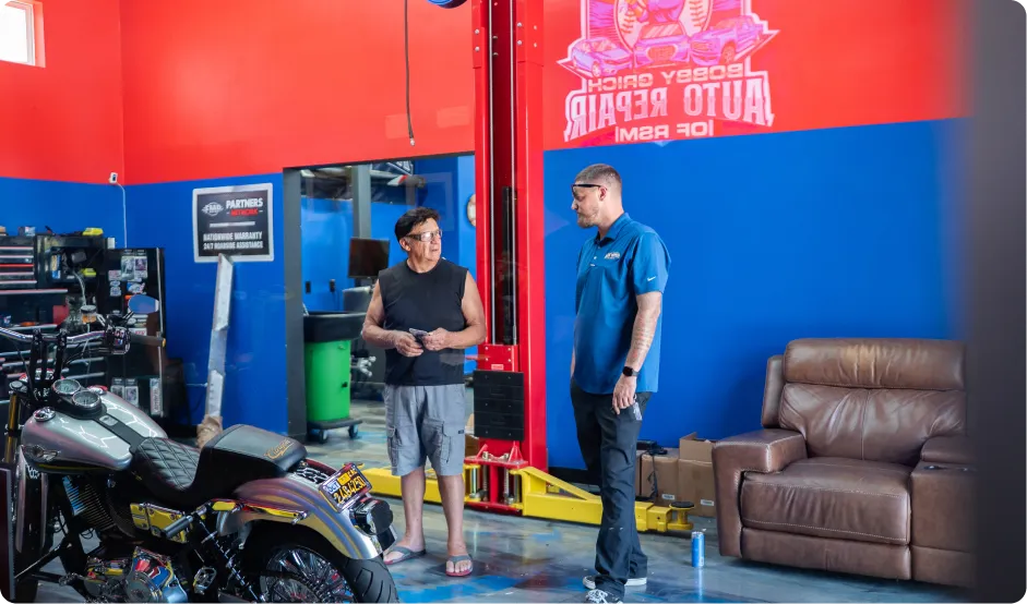 Vehicle Maintenance In Rancho Santa Margarita, CA At Bobby Grich Auto Repair. Two men stand and talk in a brightly-colored auto repair shop with red and blue walls. A motorcycle is parked nearby, and tools, a couch, and shop equipment are visible in the background.