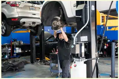 A mechanic works on exposed brakes of a car lifted in an auto repair shop Rancho Santa Margarita CA, with another car raised in the background and tools nearby.