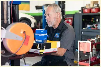 A gray-haired mechanic works in an auto repair shop in Rancho Santa Margarita, sitting at a counter and handling car parts in boxes, with tools and equipment visible in the background.