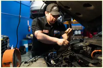 A mechanic in a black uniform and cap works on a car engine at an auto repair shop in Rancho Santa Margarita, with tools and equipment visible in the background.