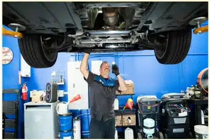 A mechanic wearing gloves works underneath a car lifted on a hydraulic lift in an auto repair shop in Rancho Santa Margarita, CA, with blue walls and various tools and equipment around.