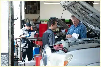 Two men stand by a car with its hood open, discussing paperwork in an auto repair shop Rancho Santa Margarita CA, while another worker in the background examines documents and equipment.
