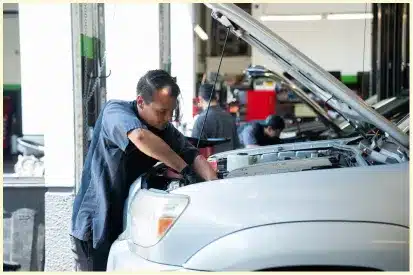 A mechanic works under the hood of a white car in an auto repair shop, one of the trusted auto repair shops in Rancho Santa Margarita, while another mechanic is busy with a different vehicle in the background.