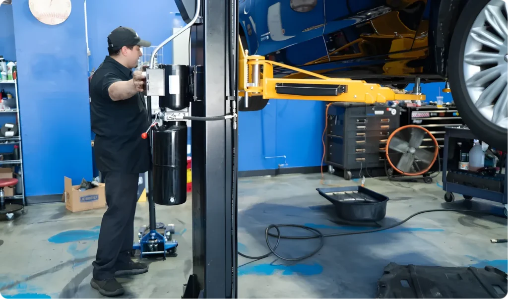 A mechanic in black uniform operates a vehicle lift in a brightly lit auto repair shop, focusing on wheel alignment repair. Part of a raised car, toolboxes, a hose, and a fluid drain pan are visible on the floor.